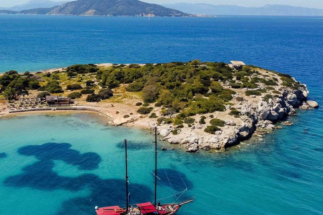 Aerial view of a red-sailed gulet anchored in turquoise water beside a small pine-covered islet with a sandy cove and beach bar