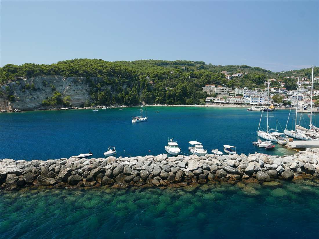 Patitiri harbour on Alonissos with moored boats, a stone breakwater and pine-covered hills above clear blue water