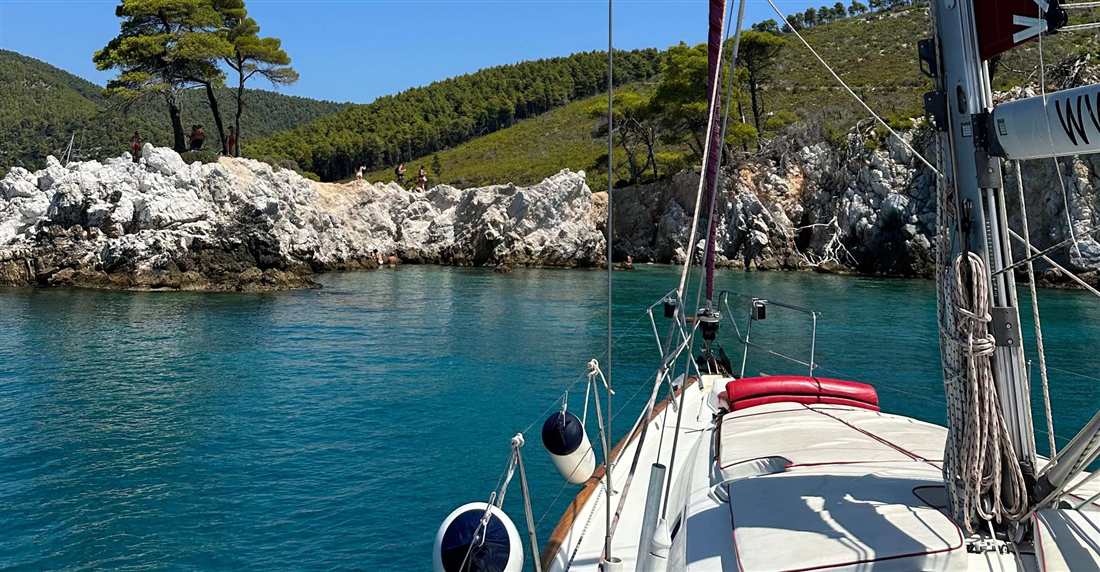 A sailing boat moored by white rocks and pine trees in a clear blue cove on a Sporades sailing cruise