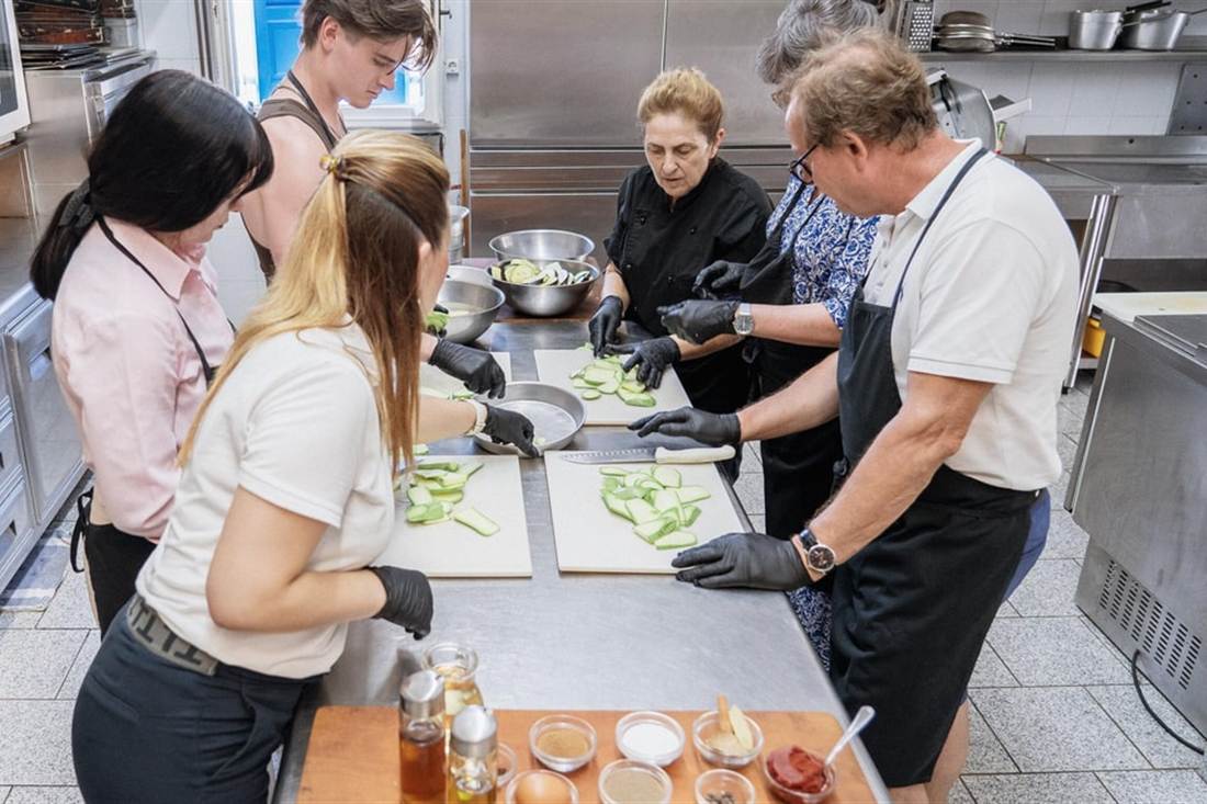 Guests in black aprons slicing vegetables at steel worktops beside the chef in a hotel kitchen during an Amorgos cooking class.