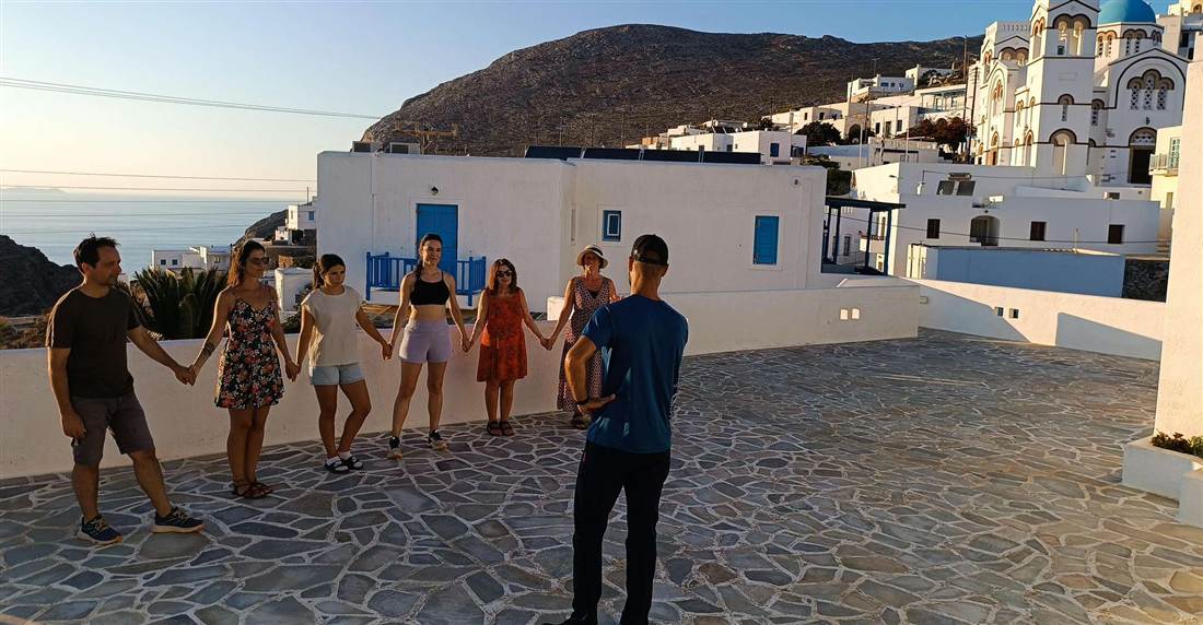 Visitors holding hands in a Greek circle dance on a whitewashed terrace on Amorgos, with a church and the sea behind them