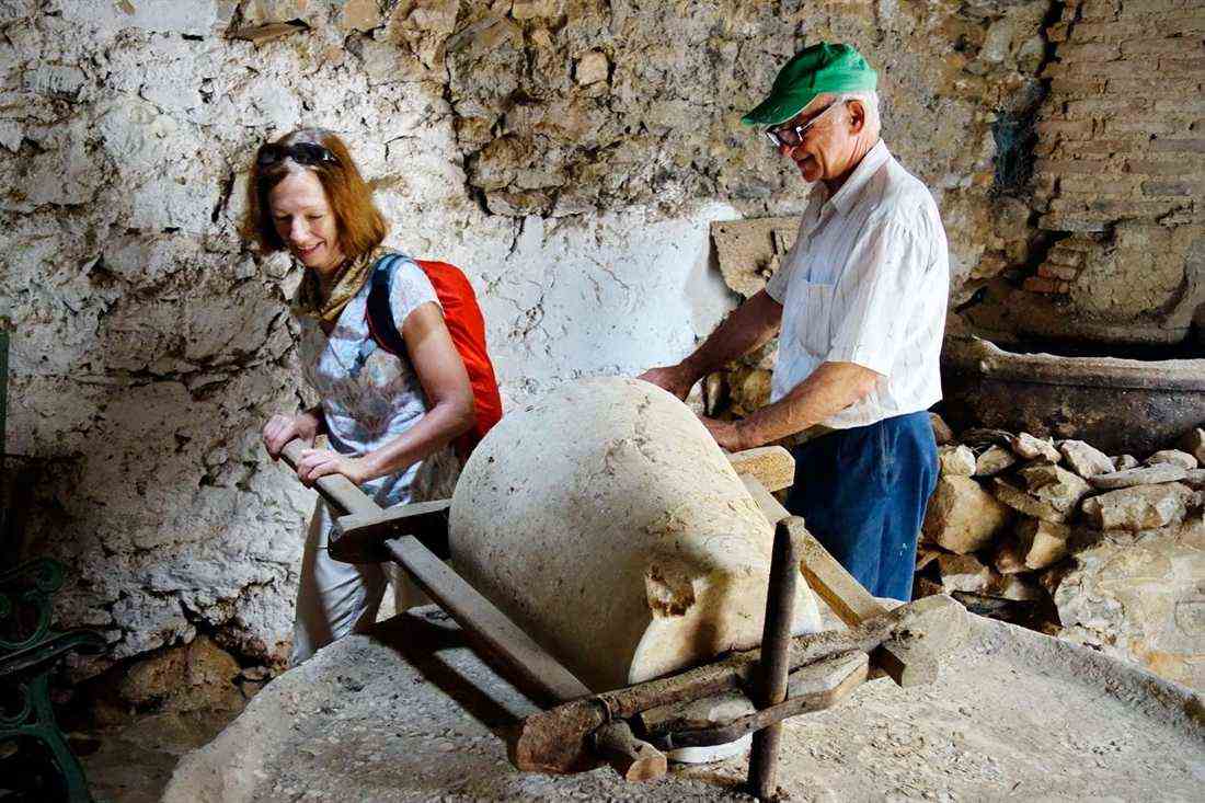 A walker and a local guide beside the stone millstone and wooden beams of an old olive press in a stone-walled room on Amorgos