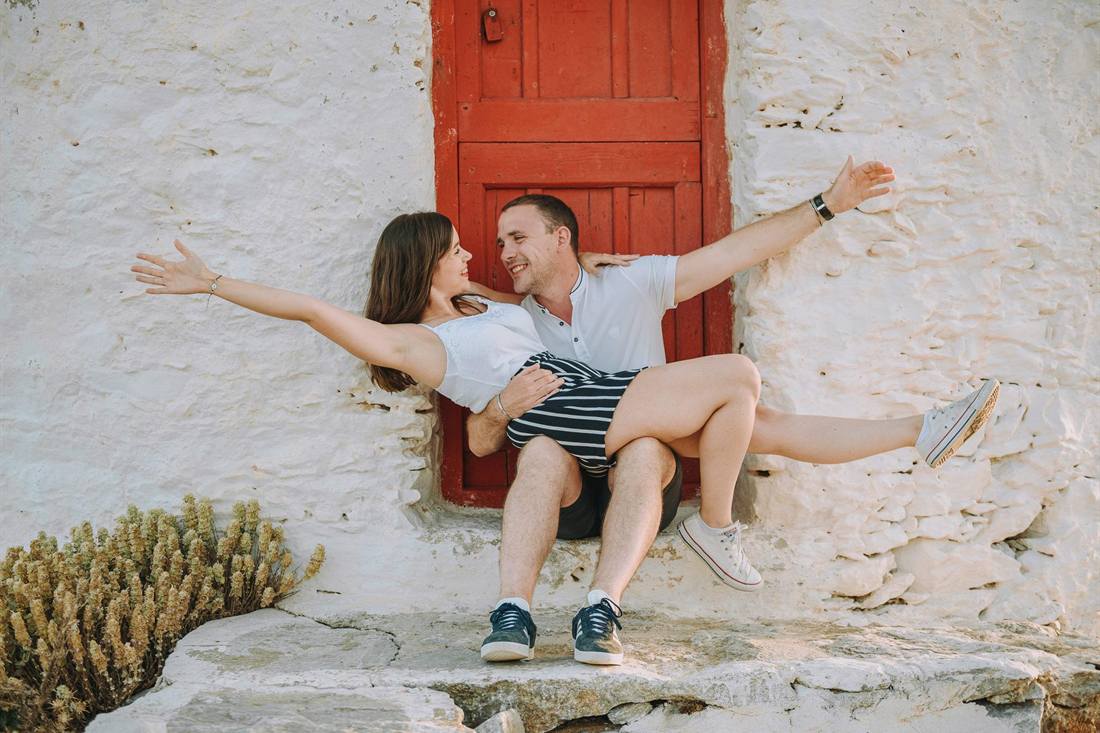 A smiling couple posing with arms spread wide on a stone step in front of a red wooden door in a whitewashed wall on Amorgos