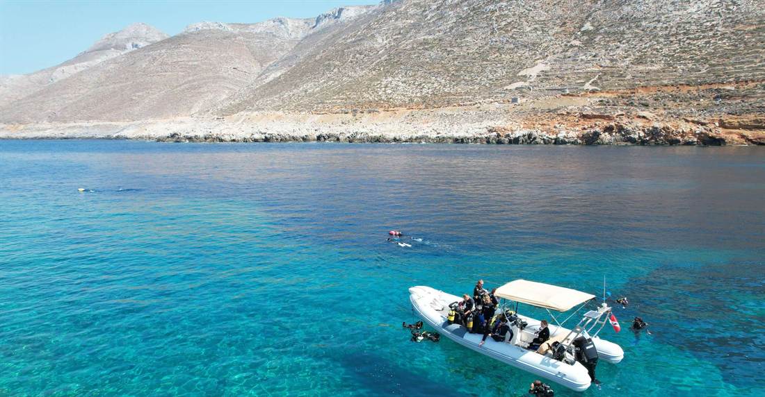 Inflatable dive boat with snorkelers and divers in clear blue water below a bare rocky Amorgos hillside