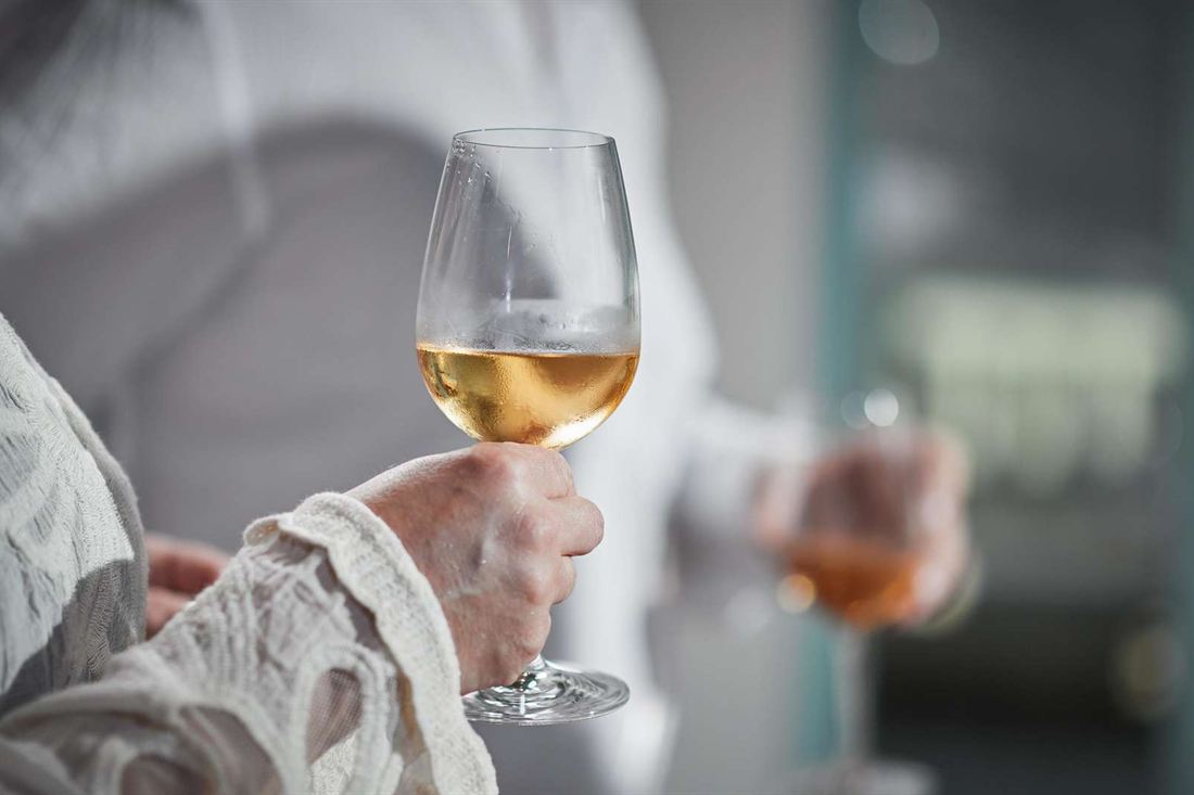 A hand holding a glass of golden wine at a tasting, with a second glass of amber wine blurred behind