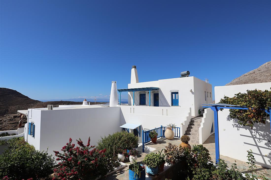 A whitewashed house with blue shutters and a blue pergola, a leafy vine climbing it, under a clear blue sky in Tholaria on Amorgos