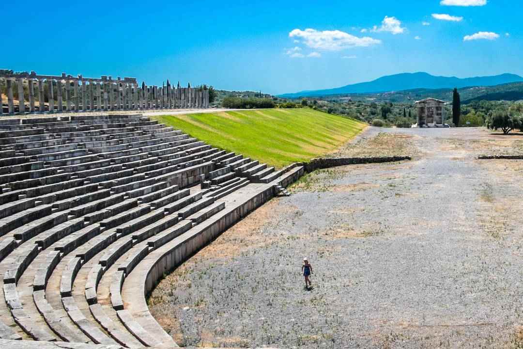 The long stone stadium of Ancient Messene with rows of seats, a colonnade at the far end and green hills under a blue sky.