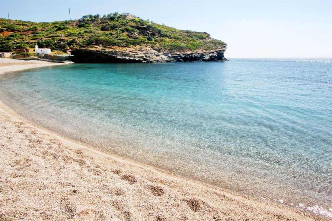 A curved pebble beach with clear turquoise water below green hills on Andros