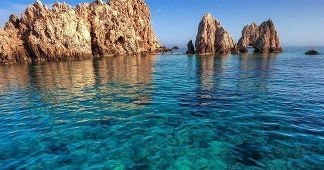Clear blue-green water over a rocky seabed beside red rock stacks on a sunny coast, seen on an Antiparos day trip from Naxos.