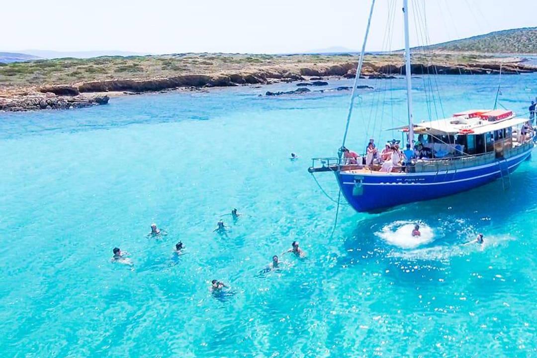 A sailing boat anchored in a turquoise lagoon off Antiparos, with swimmers in the clear water around it.