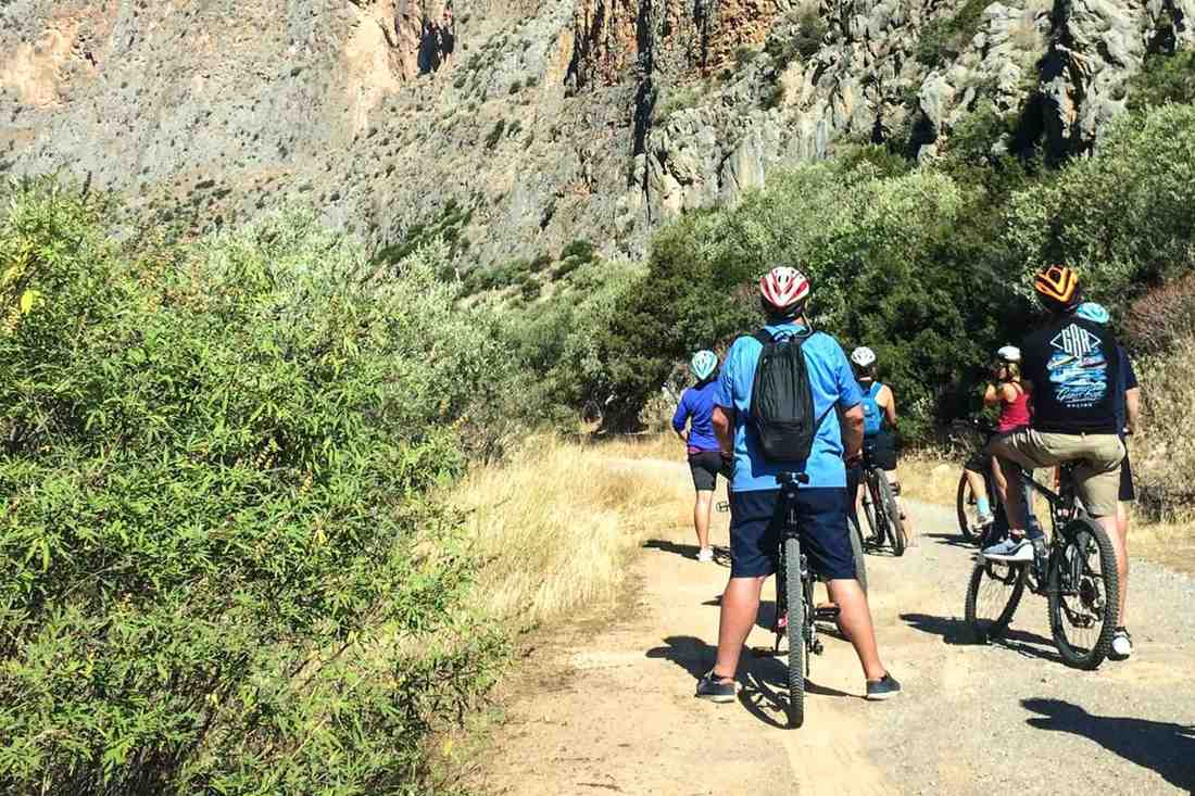 A group of cyclists in helmets stopped on a gravel track beside a tall grey rock cliff on a sunny day