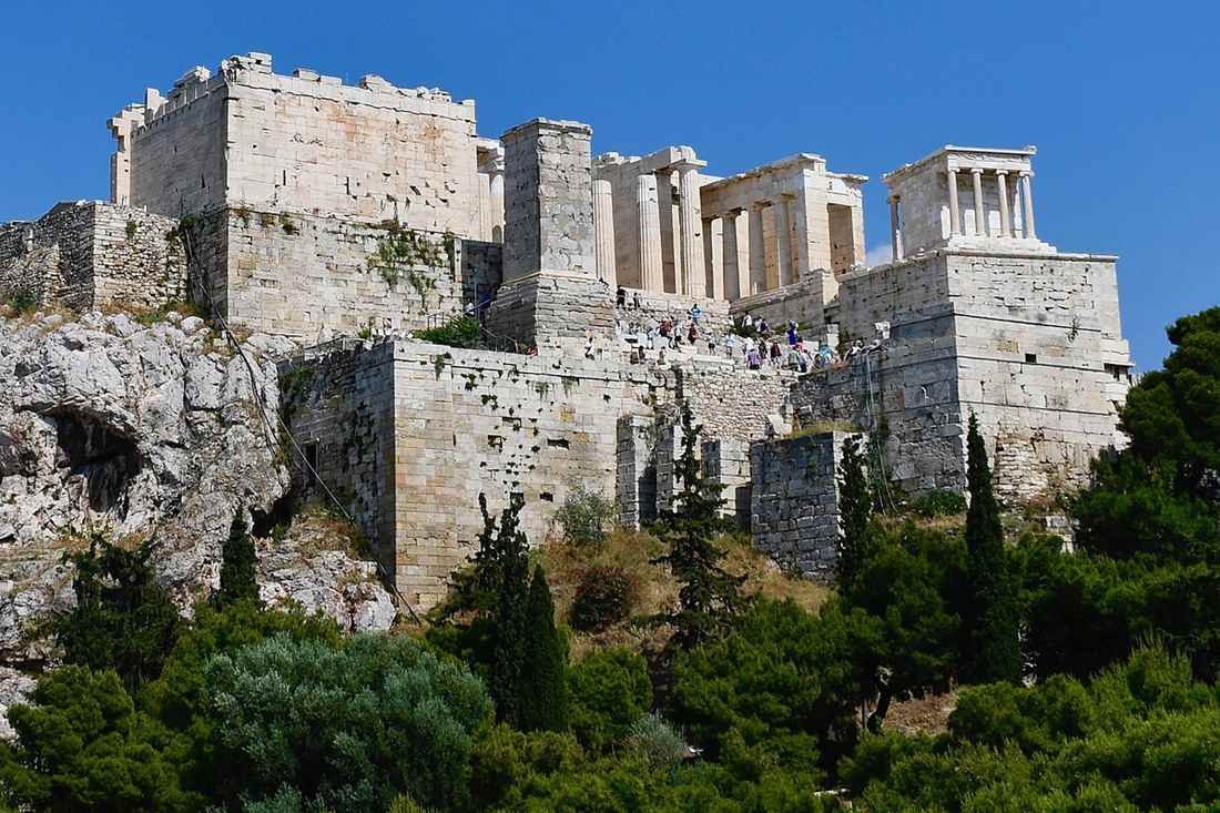 The Propylaea gateway and the small Temple of Athena Nike on the Acropolis rock above green trees under a clear blue sky