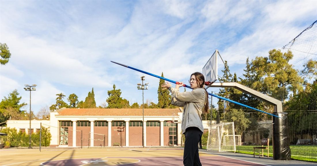 A woman throws a long javelin in a sports ground with a neoclassical building and trees behind her, during the Athens Olympic Games workout.