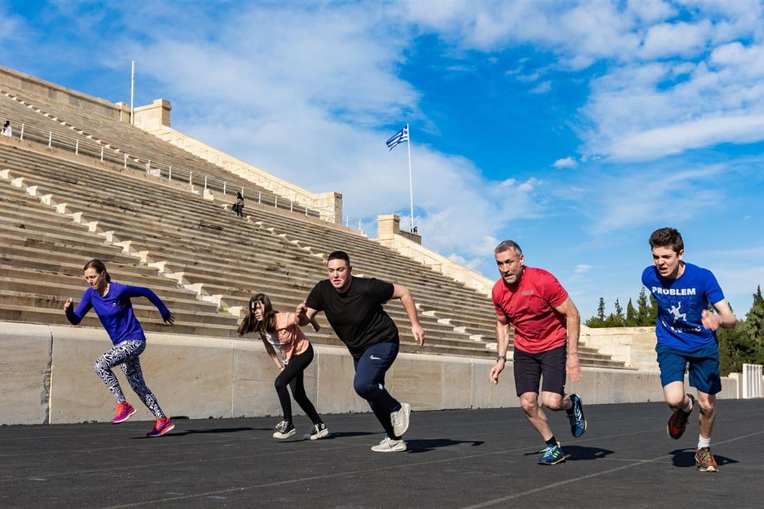 Five runners sprint along the track of the marble Panathenaic Stadium under a blue sky during the Athens Olympic Games workout.