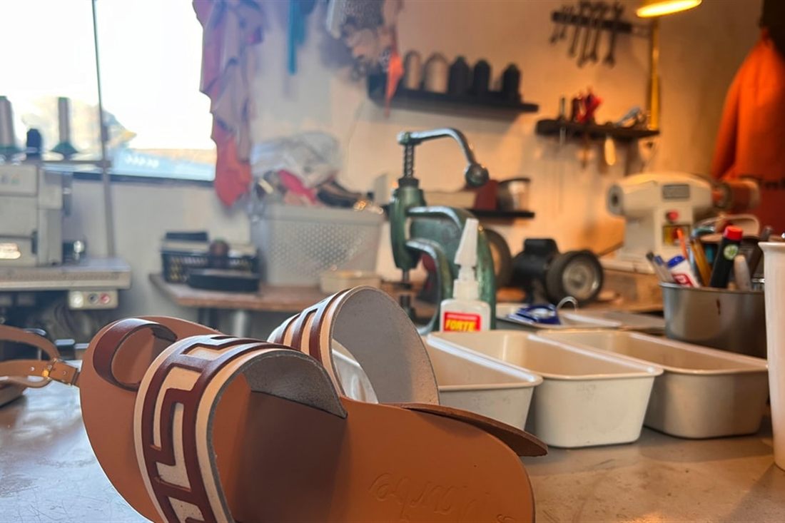 A pair of brown leather sandals with Greek key straps on a workbench beside leather tools, made at an Athens sandal workshop.