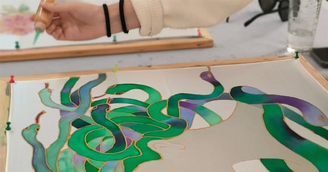 A silk square stretched on a wooden frame, painted with swirling green shapes inside fine outlines, at the Athens silk scarf workshop.