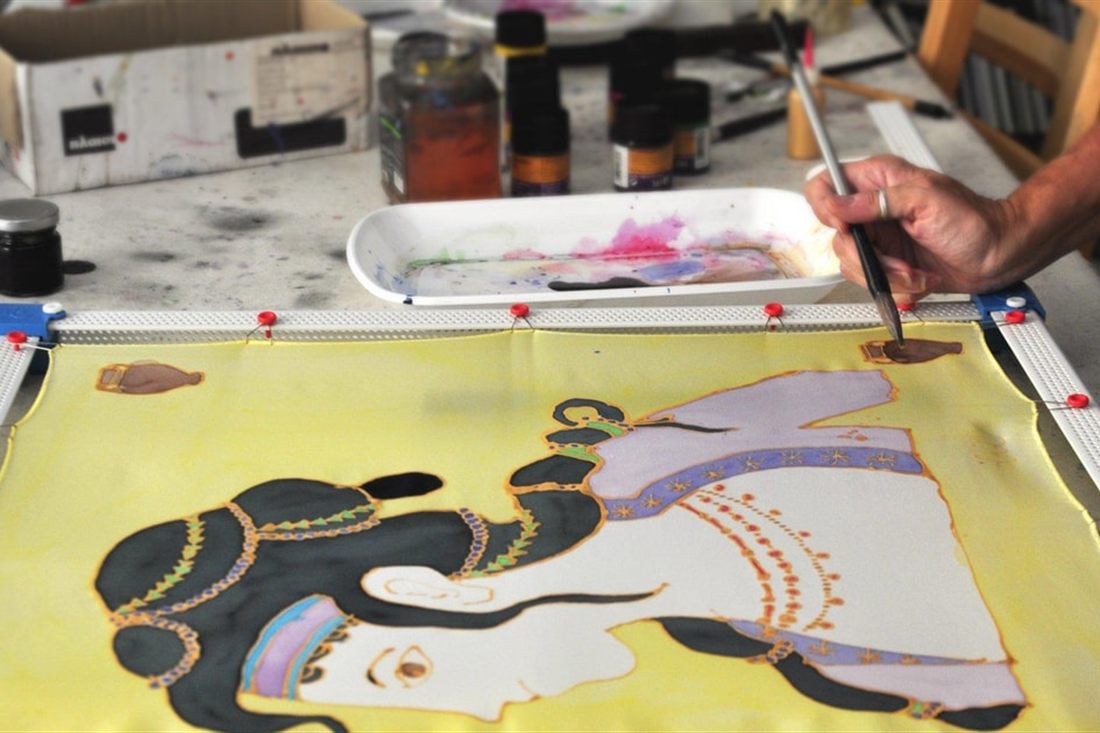 A hand paints a figure of a woman with dark hair and earrings on yellow silk stretched on a frame, at the Athens silk scarf workshop.