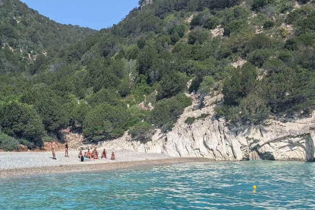 Pebble cove on Atokos seen from the water, with a few swimmers on the beach below a wooded hillside and a white limestone cliff