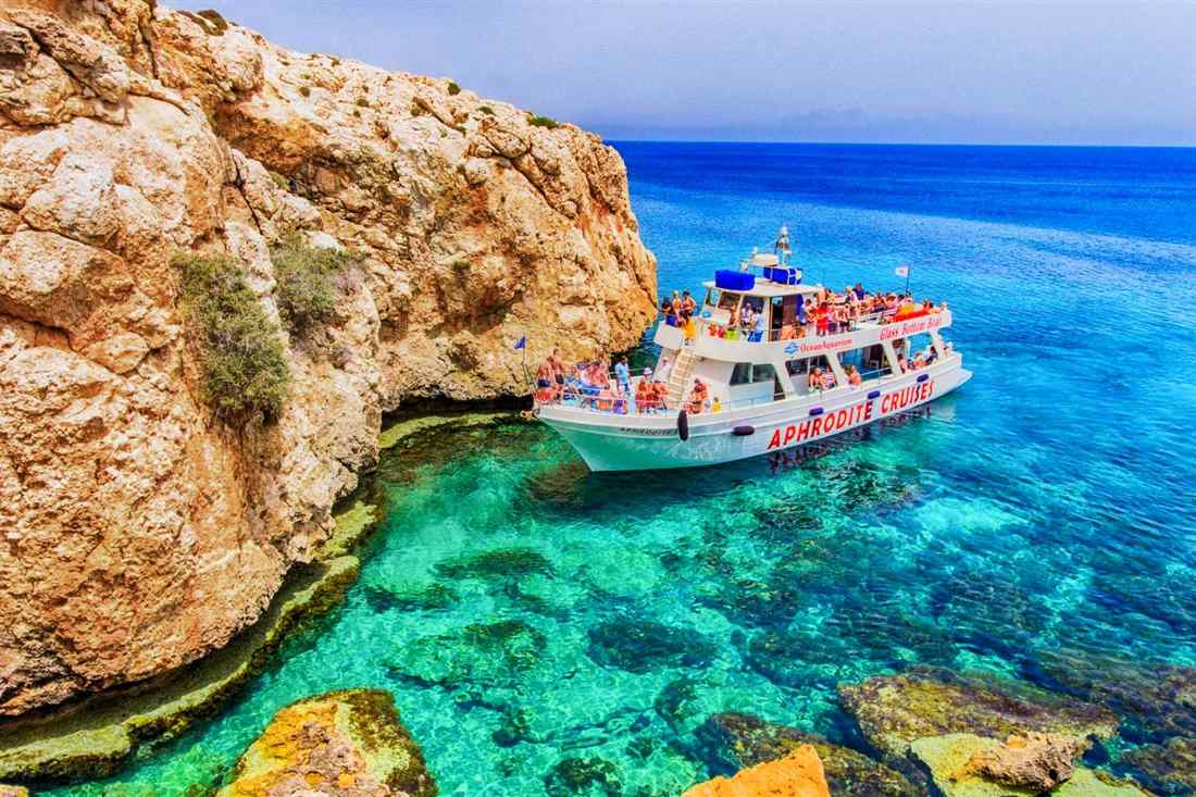 A cruise boat anchored below orange limestone cliffs near Cape Greco over clear turquoise water