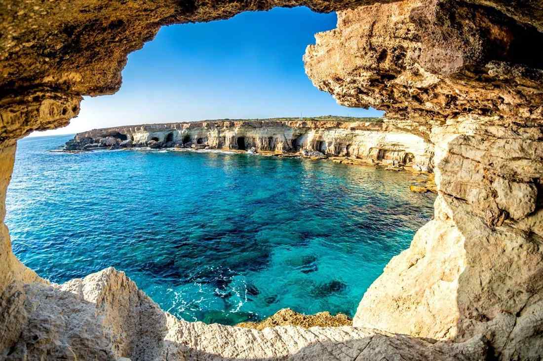 Looking out through a rocky sea cave window at turquoise water and the pale limestone sea cave cliffs of Cape Greco under a clear blue sky