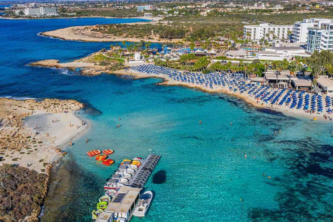 Aerial view of a sandy beach bay at Ayia Napa with rows of blue sunshades, turquoise water, a boat jetty and white hotels