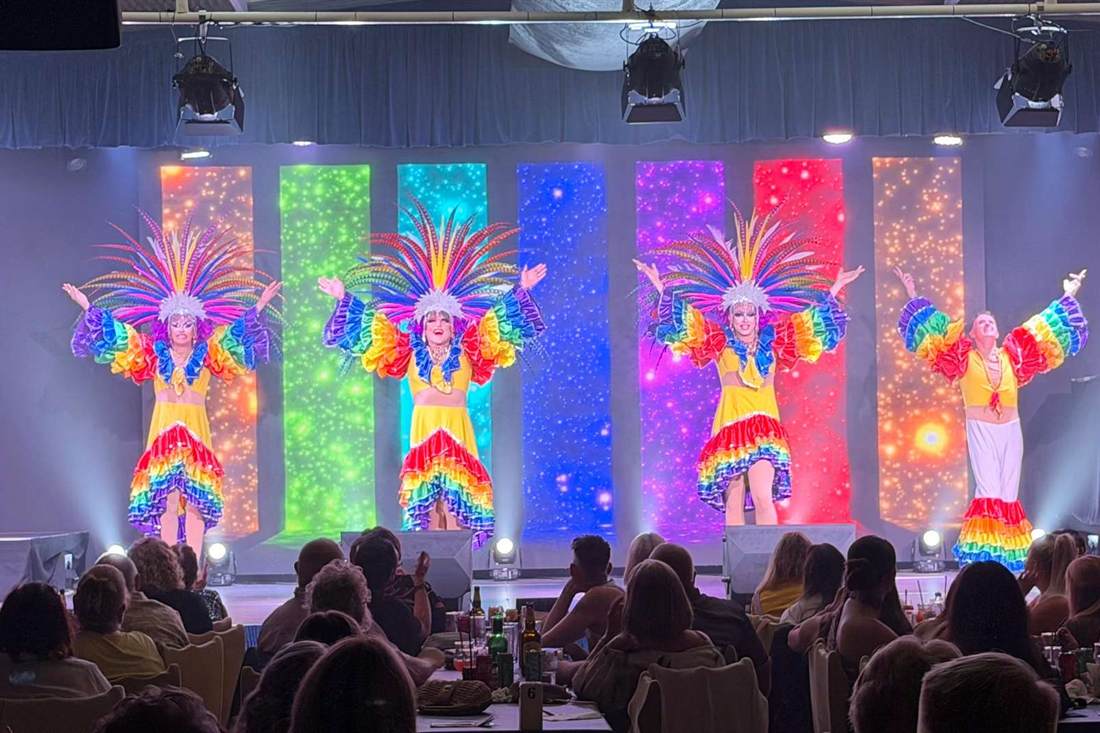 Four performers in rainbow feathered headdresses and ruffled yellow costumes on a stage with coloured lit panels, with audience tables in front