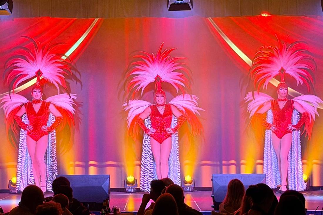 Three performers in red costumes with pink feather headdresses and zebra-striped capes standing on a stage under red and orange lights