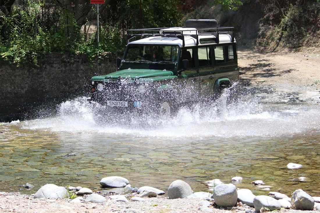 A green Land Rover Defender jeep splashing through a shallow stream beside a dirt track