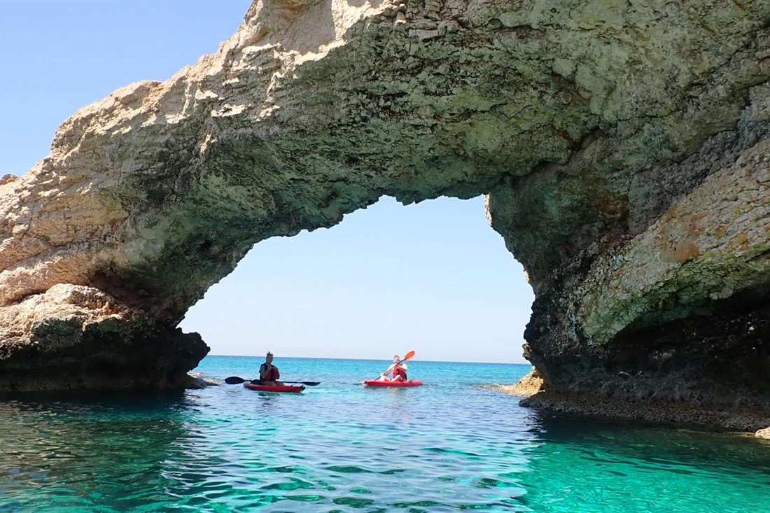Two kayakers in red kayaks paddle on turquoise water beneath a large limestone sea arch on the Cape Greco coast near Ayia Napa