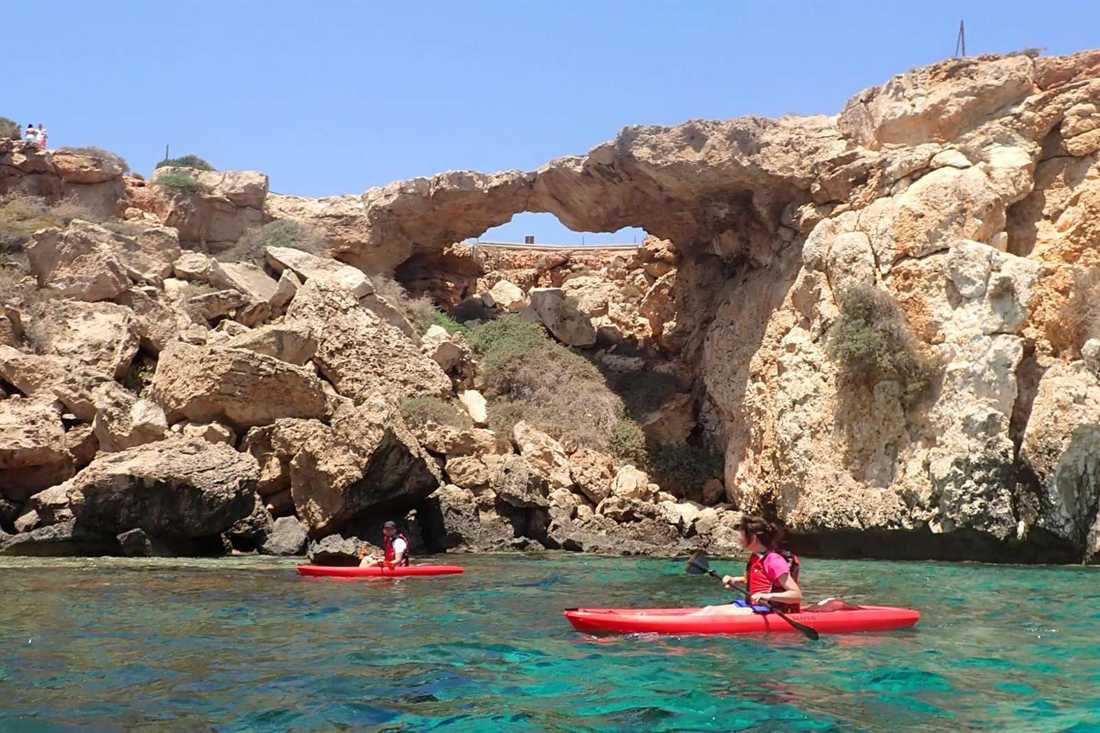 Two kayakers in red kayaks and life jackets paddle on clear turquoise water below a rocky Cape Greco cliff with a natural stone bridge