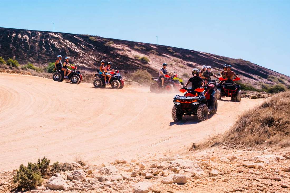 Riders on quad bikes taking a bend on a dusty off-road track past a dry hillside under a clear blue sky