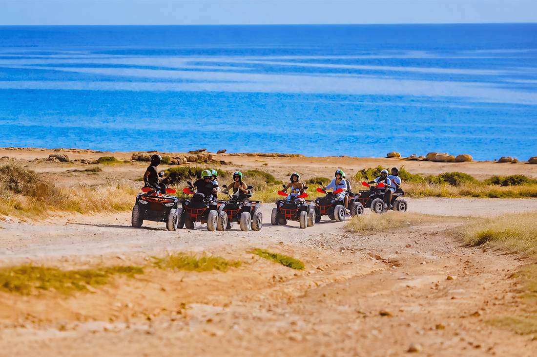 Row of quad bikes with helmeted riders on a coastal dirt track above a deep blue sea
