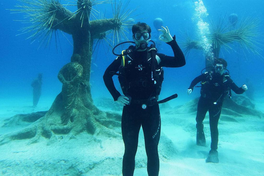 Two scuba divers in black wetsuits hover over white sand beside tree-shaped sculptures with spiky branches in clear blue water at the MUSAN underwater museum, one diver making the OK sign