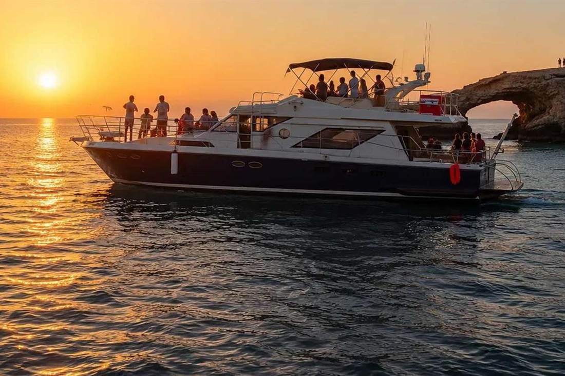 A motor yacht with passengers on its decks cruising at sunset off the Ayia Napa coast, the orange sun low over the sea and a rock arch on the cliff at right