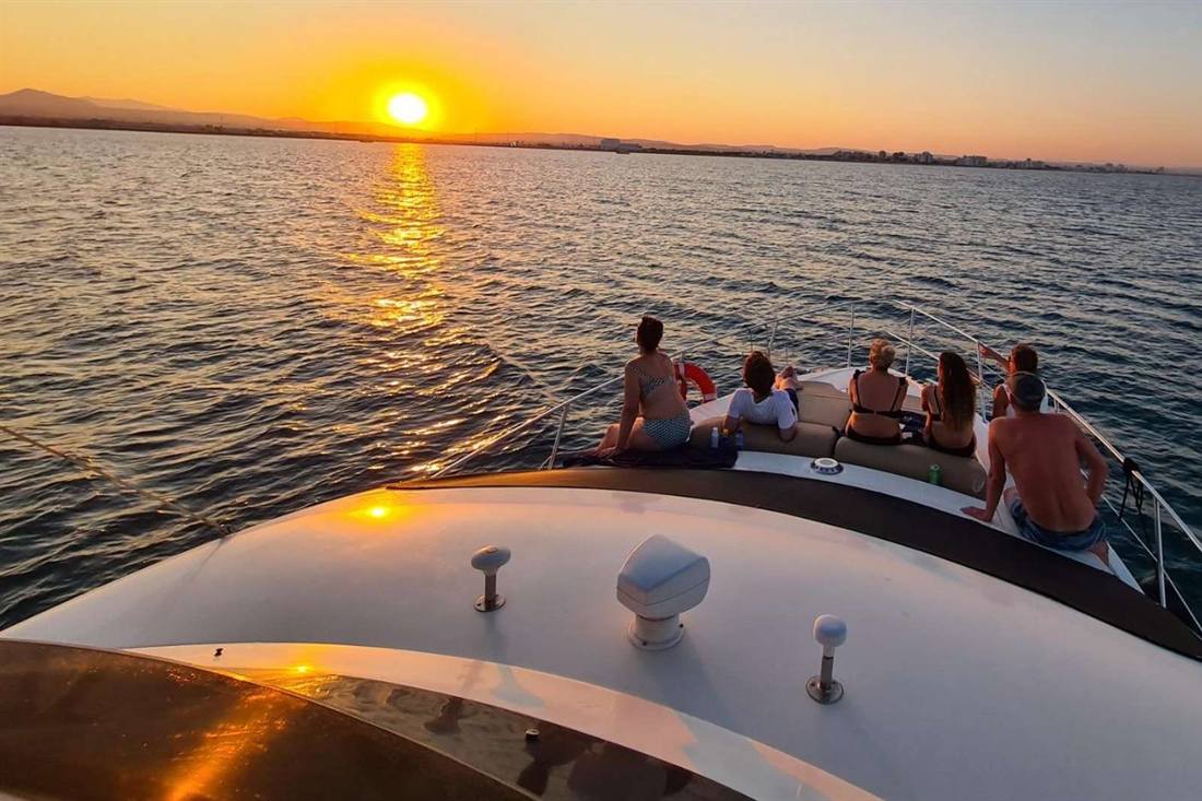 A group of passengers sitting on the bow of a yacht and watching the orange sun set over the sea, with a low coastline on the horizon