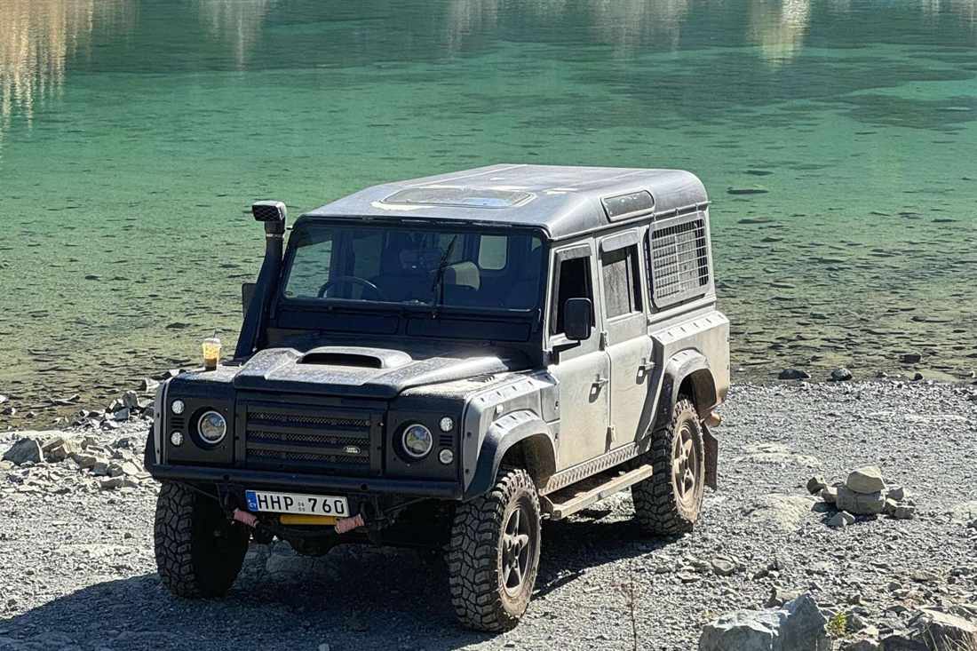 A Land Rover jeep parked on a pebble shore beside the green water of a mountain lagoon