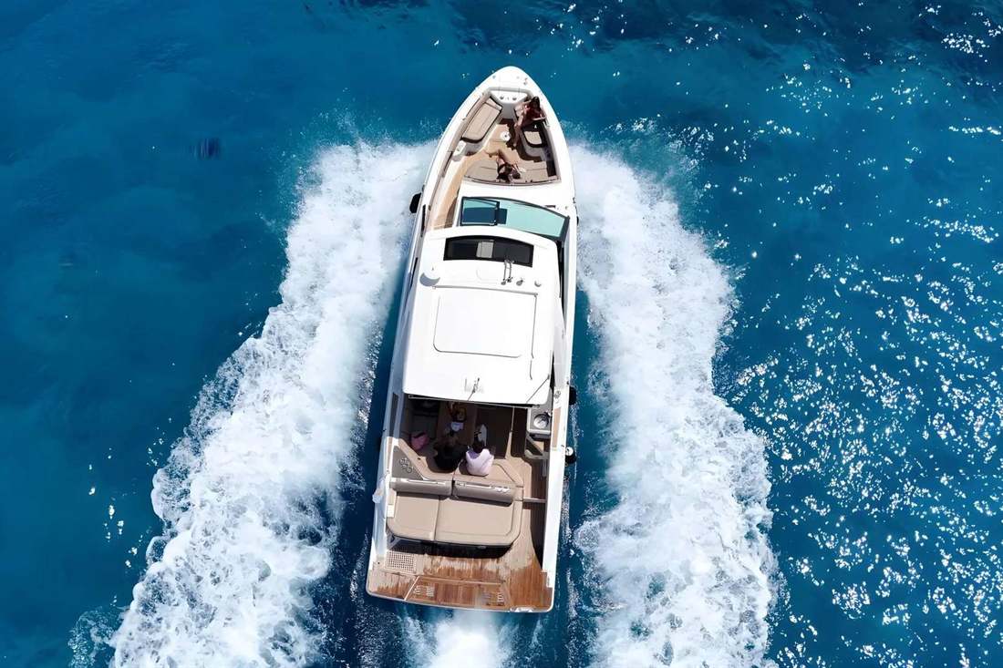 Aerial view of a white charter yacht cruising on deep blue water with a white wake and guests seated on the bow and rear deck
