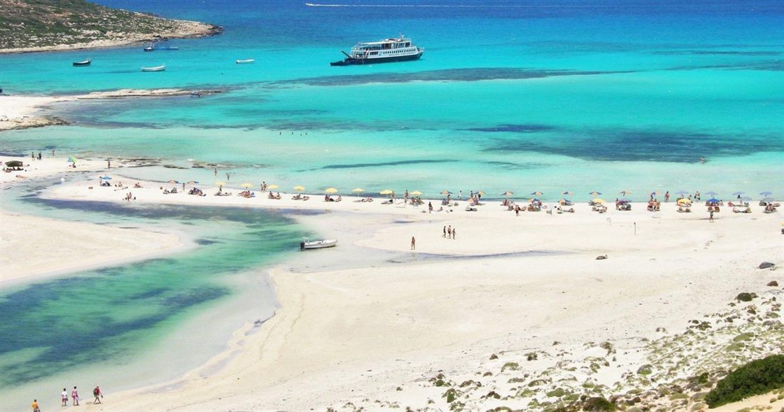 A white sandy beach with sunshades along shallow turquoise water, with a large boat moored offshore, on a Balos day trip from Rethymno.