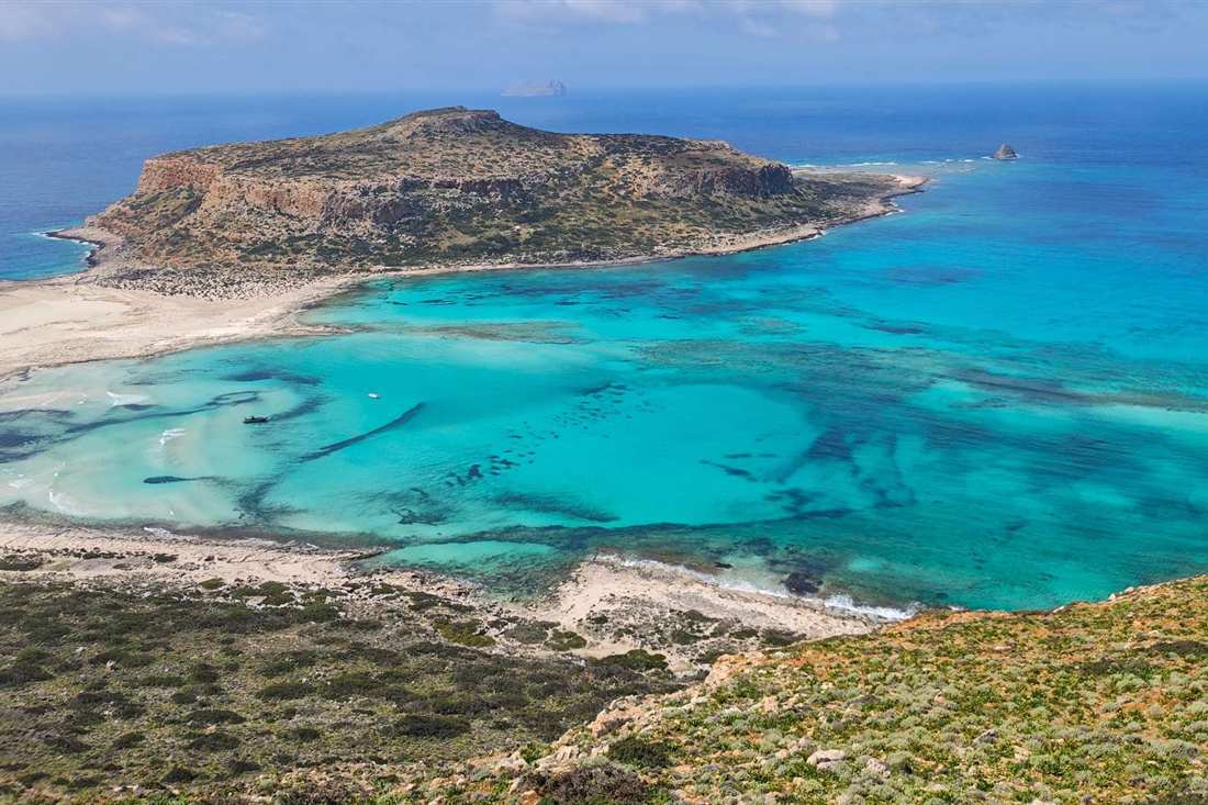 Balos Lagoon seen from the hillside above, with turquoise shallows, pale sand and a flat-topped rocky headland beyond
