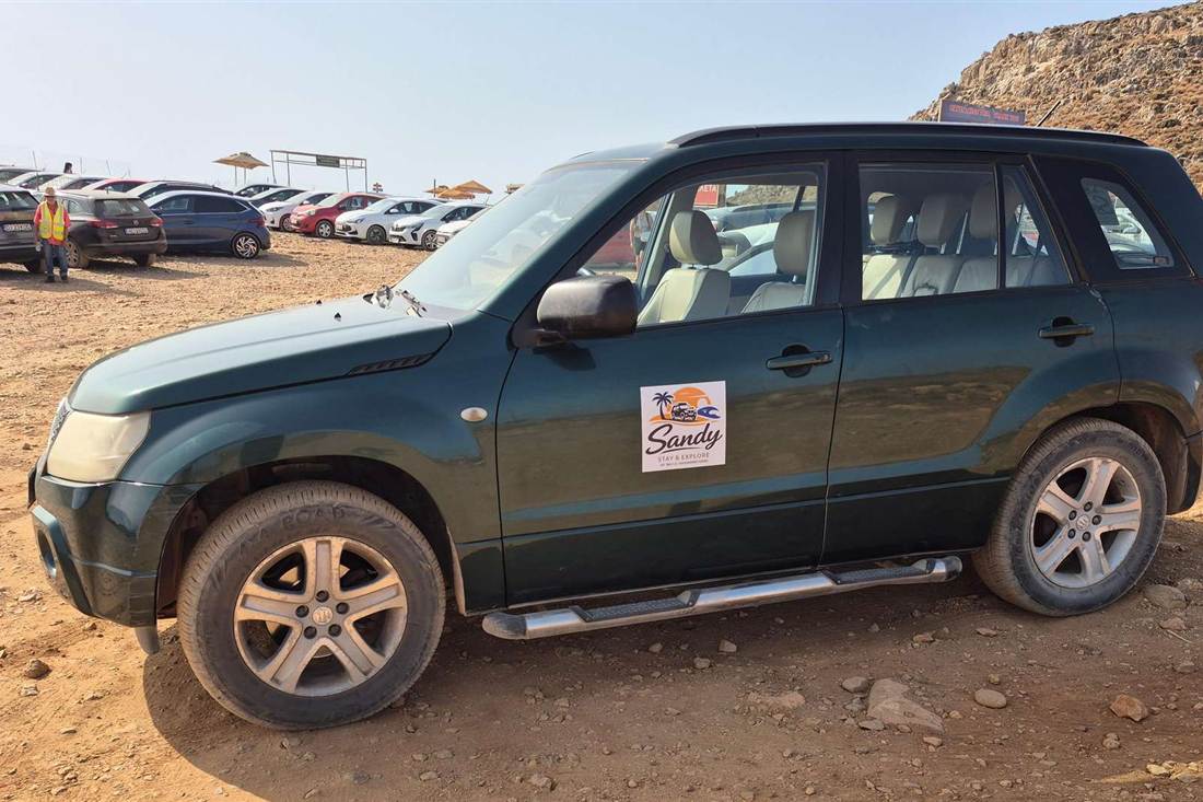 A dark green 4x4 jeep with a Sandy sticker on its door parked on bare dirt beside rows of parked cars below a rocky hill