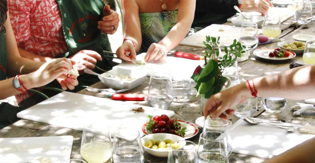 Guests preparing food with fresh herbs at a shared outdoor table during a cooking class.