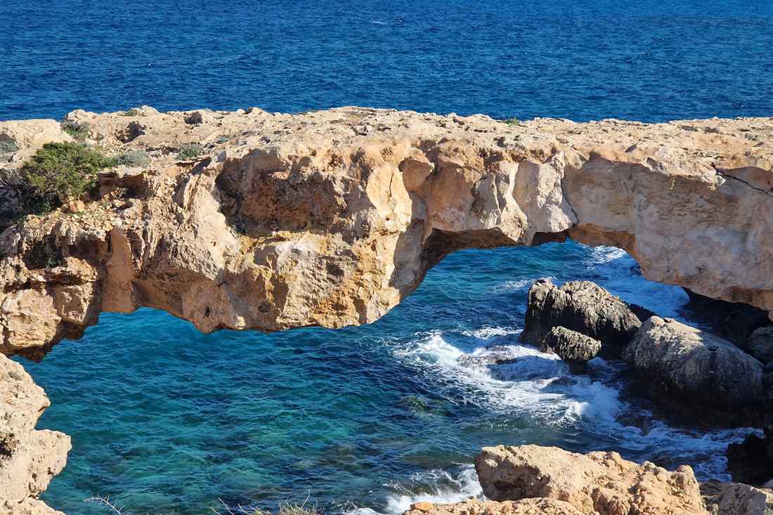 Natural sand-coloured rock arch over deep blue sea with waves breaking on dark rocks beneath it