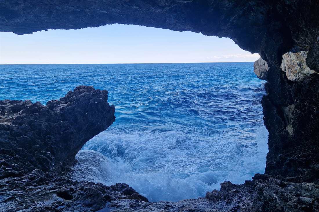View from inside a dark sea cave out to deep blue waves breaking on rocks under a pale sky