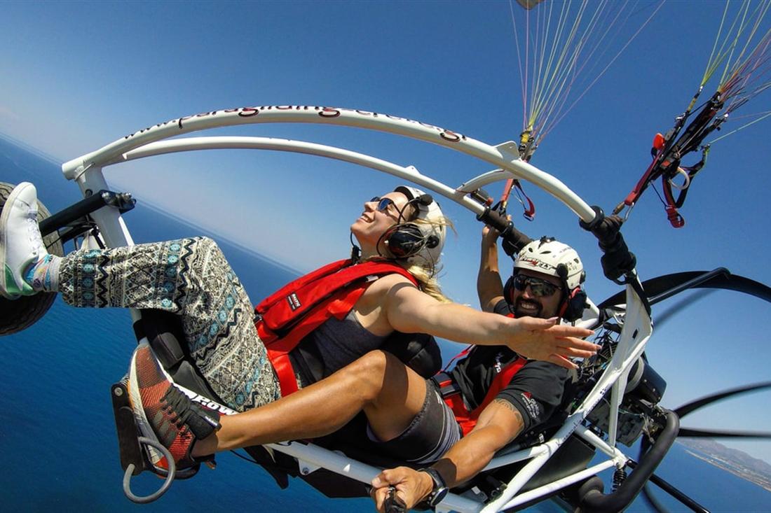 A pilot and a passenger in helmets fly in an open paratrike under its wing above the blue sea on Chania paratriking.