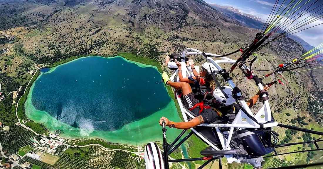 A paratrike with a pilot and passenger flies under its wing high above a round green lake ringed by hills, seen on Chania paratriking.