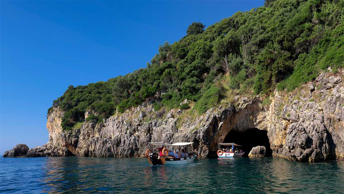 Two tour boats at the entrance of a sea cave below green cliffs at Paleokastritsa, the classic stop of a Corfu cave tour.