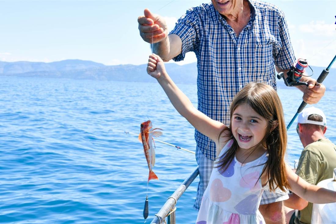 A smiling girl holds up a small red fish on a line as a man with a fishing rod stands behind her, on a Corfu fishing trip.