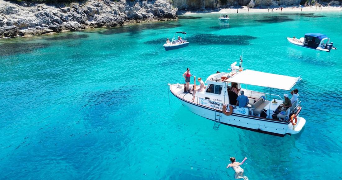 A white fishing boat anchored in a turquoise cove below a wooded rocky shore, with a swimmer in the water, on a Corfu fishing trip.