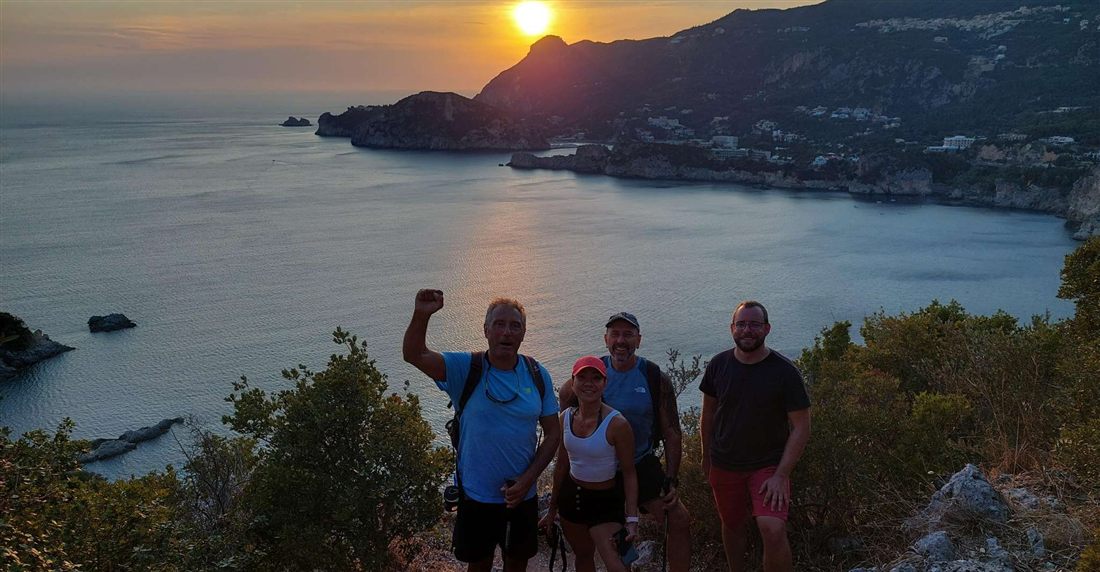 Four hikers on a hill path above the bays of Paleokastritsa on Corfu as the sun sets over the headland