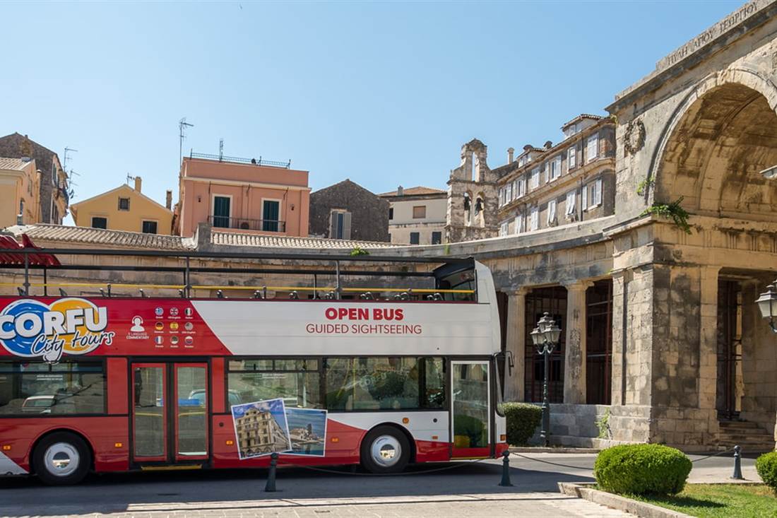 A red and white Corfu open-top sightseeing bus parked in front of a stone arcade and old buildings in Corfu Town, used for Corfu hop on hop off.